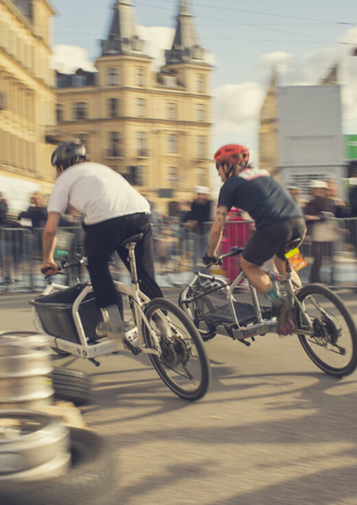 Two riders racing cargo bikes past a crowd on a city street